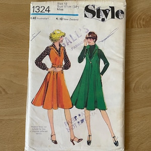 May include: A vintage sewing pattern envelope for a pinafore dress. The pattern is for size 12, bust 87 cm. The envelope features two illustrations of women wearing the dress, one in orange and one in green. The pattern is from Gall's Fabrics and is called "Style".