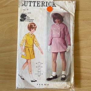 Könnte beinhalten: Vintage-Schnittmuster für ein Mädchenkleid und eine Jacke. Das Schnittmuster ist von Butterick und in Größe 8 mit einem Brustumfang von 26 Zoll. Das Schnittmuster beinhaltet ein gelbes Kleid mit weißem Kragen und eine rosa Jacke mit weißem Kragen.