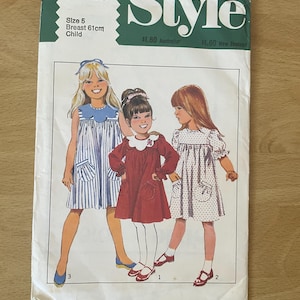May include: A vintage sewing pattern for children's dresses. The pattern is for sizes 5, with a 61 cm chest. The pattern includes three different styles of dresses, one with a striped bodice and skirt, one with a red bodice and skirt, and one with a white bodice and a polka dot skirt. The pattern is from Style magazine and is priced at $1.60 in Australia and New Zealand.