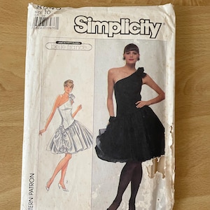 May include: Simplicity sewing pattern envelope for a black one-shoulder dress with a ruffle detail and a white one-shoulder dress with a bow detail. The pattern is size 10 and is for a Belle France design.