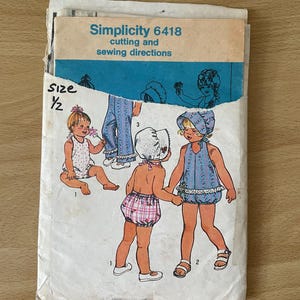 Op de afbeelding: Simplicity naaipatroon nummer 6418 voor kinderkleding. Het patroon bevat instructies voor het knippen en naaien. Het patroon toont drie verschillende outfits voor meisjes, waaronder een blauw-witte jurk met een bijpassende muts, een roze-witte gingham bloomer met een wit topje en een witte romper met een witte muts.