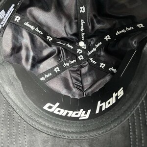 May include: The interior of a black baseball cap featuring a smooth, dark satin lining. Black fabric tapes printed with the white text "dandy hats" and a logo cross the crown. A black sweatband with the white text "dandy hats" is visible.