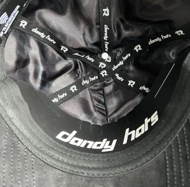 May include: The interior of a black baseball cap featuring a smooth, dark satin lining. Black fabric tapes printed with the white text "dandy hats" and a logo cross the crown. A black sweatband with the white text "dandy hats" is visible.