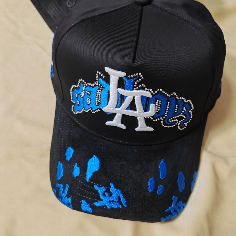 May include: A black baseball cap featuring a white and blue embroidered logo that reads "sad boys" with a large white "LA" emblem in the center. The brim is decorated with blue embroidered splatter patterns, and the logo is outlined with small silver studs.