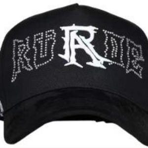 May include: Black baseball cap with the word "RURE" in white, embellished with rhinestones. The brim is black, and the cap has a curved visor. The cap is a stylish accessory.
