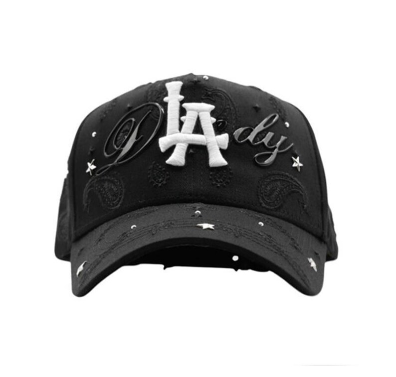 May include: Black baseball cap with the text "LA dy" in white and silver. The cap has a paisley pattern and star-shaped embellishments. The curved peak and structured crown give it a classic baseball cap appearance.