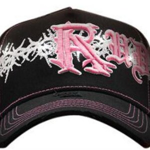 May include: Black baseball cap with a curved brim, featuring pink embroidered lettering and white decorative accents. The cap has a pink stitched brim and a structured design.