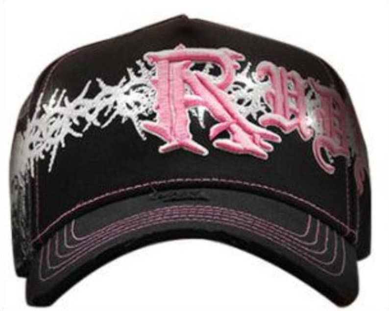 May include: Black baseball cap with a curved brim, featuring pink embroidered lettering and white decorative accents. The cap has a pink stitched brim and a structured design.