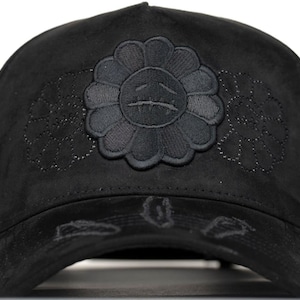May include: A black baseball cap featuring a central embroidered flower motif with a sad face expression. The cap is made of a soft, suede-like material and includes additional subtle, tonal floral embroidery on the sides and distressed detailing on the brim.