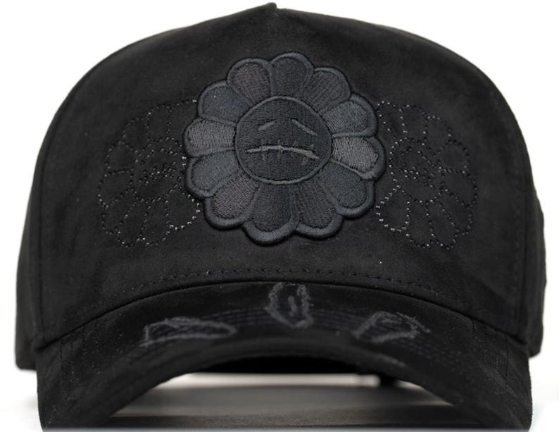 May include: A black baseball cap featuring a central embroidered flower motif with a sad face expression. The cap is made of a soft, suede-like material and includes additional subtle, tonal floral embroidery on the sides and distressed detailing on the brim.