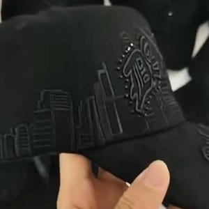 K&ouml;nnte beinhalten: Eine schwarze Baseballkappe mit einer gestickten Stadtlandschaft auf dem Seitenteil und einem stilisierten Logodetail. Die Kappe hat eine strukturierte Krone und einen gebogenen Schirm mit durchgehenden schwarzen N&auml;hten und Verzierungen.