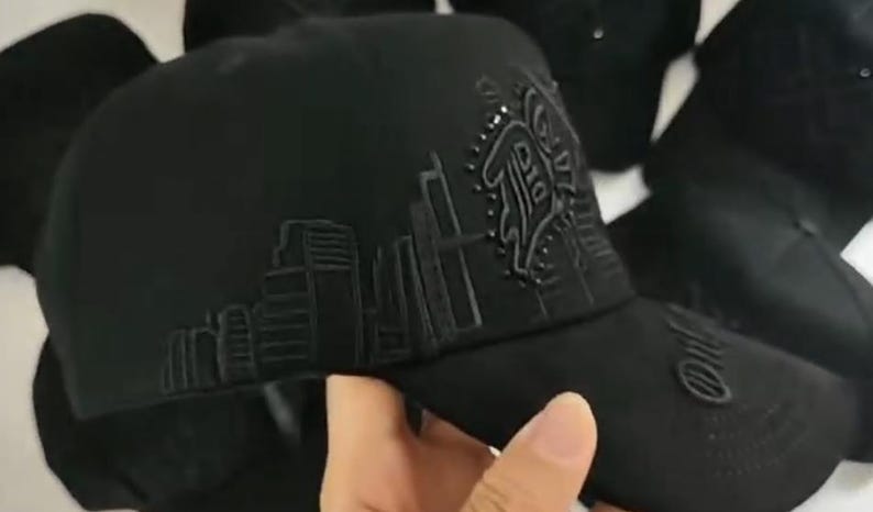K&ouml;nnte beinhalten: Eine schwarze Baseballkappe mit einer gestickten Stadtlandschaft auf dem Seitenteil und einem stilisierten Logodetail. Die Kappe hat eine strukturierte Krone und einen gebogenen Schirm mit durchgehenden schwarzen N&auml;hten und Verzierungen.