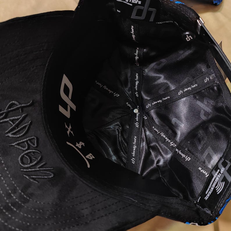 May include: A black baseball cap viewed from the inside, featuring a black satin lining with repeating white dandy hats branding on the interior seams. The inner sweatband displays the text BADBOY in black embroidery and a white logo graphic.