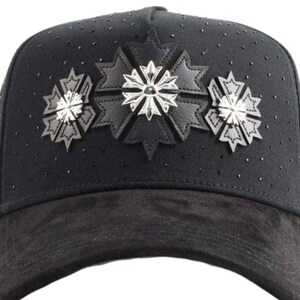 May include: A black baseball cap featuring a suede-textured brim and a crown adorned with small black studs. The front panel displays three black leather star-shaped appliqu&eacute;s, each centered with a polished silver-toned metallic cross emblem.