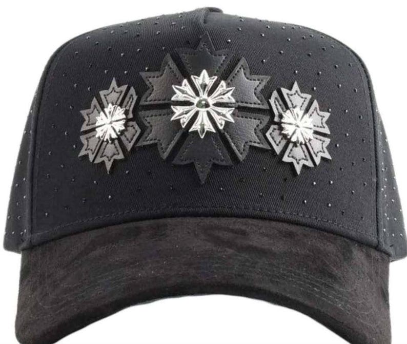 May include: A black baseball cap featuring a suede-textured brim and a crown adorned with small black studs. The front panel displays three black leather star-shaped appliqu&eacute;s, each centered with a polished silver-toned metallic cross emblem.