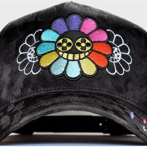 May include: A black velvet baseball cap featuring a large, central embroidered flower with multi-colored petals and a smiling face with four-leaf clover eyes. Two smaller, white-outlined flower silhouettes flank the central design.