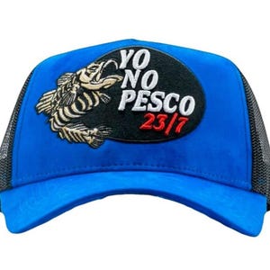 May include: A blue trucker cap with a black mesh back. The front has a black oval patch with the words "YO NO PESCO 23/7" in white and red. An embroidered fish design is on the left side of the patch.
