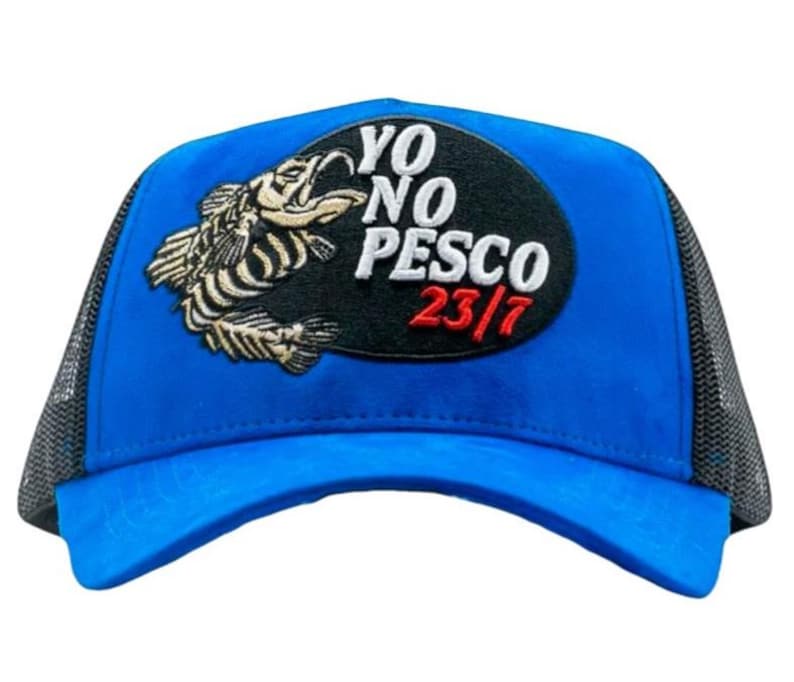 May include: A blue trucker cap with a black mesh back. The front has a black oval patch with the words "YO NO PESCO 23/7" in white and red. An embroidered fish design is on the left side of the patch.