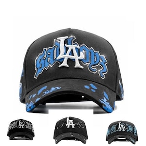 May include: Black baseball cap with the text "LA BADDS" in white and blue. The cap features a curved brim and a decorative design on the front. The hat is a stylish accessory.