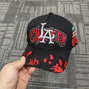May include: A black baseball cap featuring the text "chLAry" in red and white lettering with rhinestone accents. The brim displays a red splatter pattern, and the side of the cap includes a graphic of two red cherries with green stems and leaves.