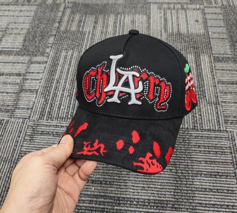 May include: A black baseball cap featuring the text "chLAry" in red and white lettering with rhinestone accents. The brim displays a red splatter pattern, and the side of the cap includes a graphic of two red cherries with green stems and leaves.
