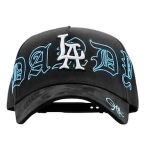 May include: Black baseball cap with the text "LA DID" embroidered in white and light blue. The cap has a curved brim and a structured crown. The design features stylized lettering.