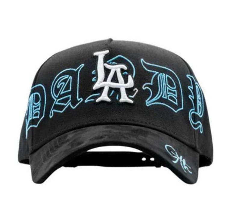 May include: Black baseball cap with the text "LA DID" embroidered in white and light blue. The cap has a curved brim and a structured crown. The design features stylized lettering.