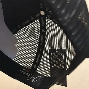 May include: Black trucker hat with a mesh back and a curved peak. The interior has a white lining and a tag with the text "OSCAR ORTIZ". The hat has a snap closure and a label with a signature.