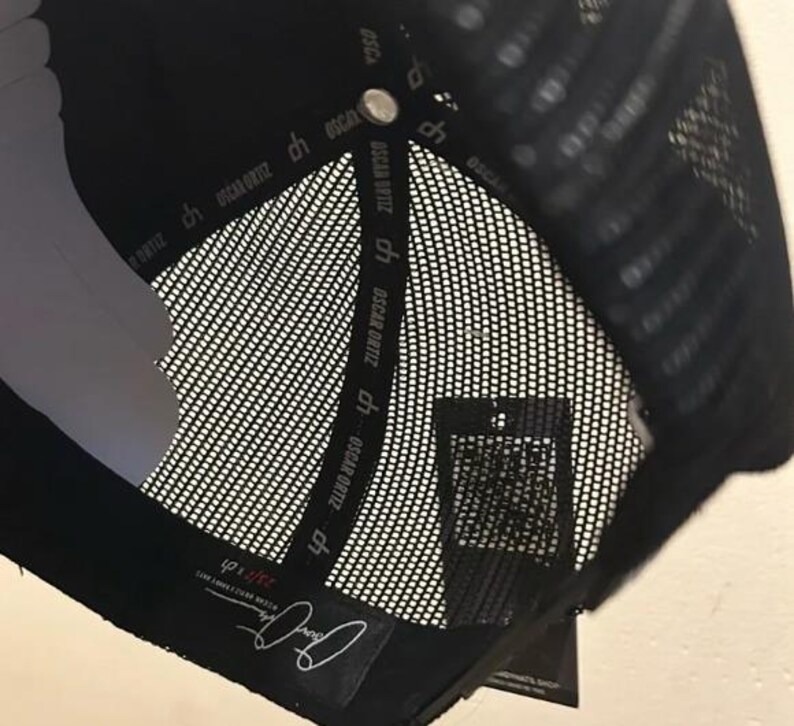 May include: Black trucker hat with a mesh back and a curved peak. The interior has a white lining and a tag with the text "OSCAR ORTIZ". The hat has a snap closure and a label with a signature.