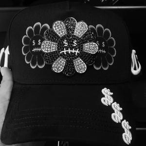 May include: A black baseball cap featuring a central flower emblem encrusted with black and clear crystals. The flower has a dollar sign for eyes and a stitched mouth. Two embroidered flower outlines flank the center, and dollar signs adorn the brim.