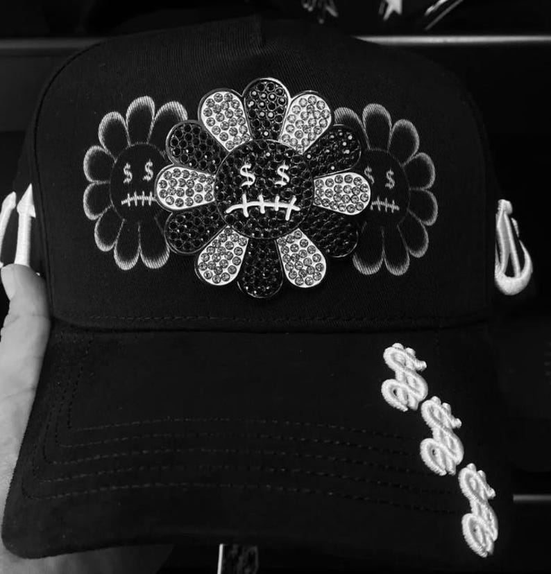 May include: A black baseball cap featuring a central flower emblem encrusted with black and clear crystals. The flower has a dollar sign for eyes and a stitched mouth. Two embroidered flower outlines flank the center, and dollar signs adorn the brim.