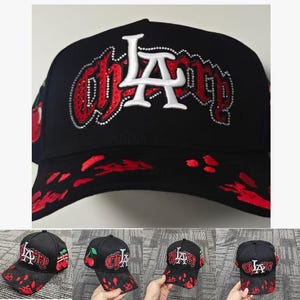 May include: A black baseball cap featuring the white and red embroidered text "CHERRY" with a large white "LA" logo overlay. The design includes red rhinestone accents, a red cherry graphic on the side, and a red paint splatter pattern on the brim.