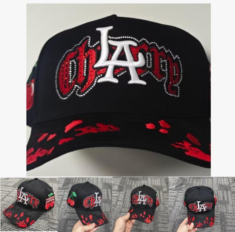 May include: A black baseball cap featuring the white and red embroidered text "CHERRY" with a large white "LA" logo overlay. The design includes red rhinestone accents, a red cherry graphic on the side, and a red paint splatter pattern on the brim.