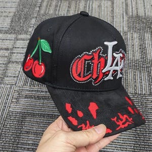 May include: A black baseball cap featuring an embroidered red cherry graphic on the side. The front displays the text CHLA in red and white lettering outlined with small silver studs. The brim has a red splatter pattern across the black fabric.
