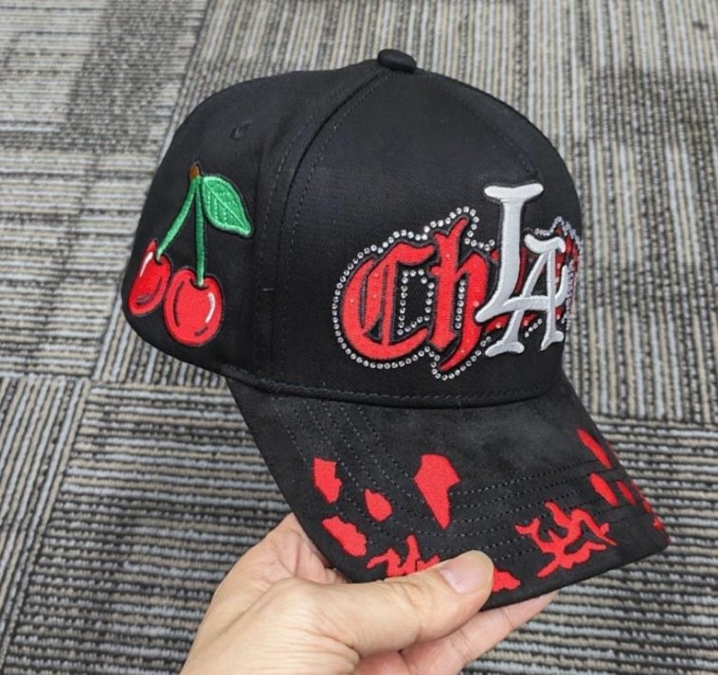 May include: A black baseball cap featuring an embroidered red cherry graphic on the side. The front displays the text CHLA in red and white lettering outlined with small silver studs. The brim has a red splatter pattern across the black fabric.