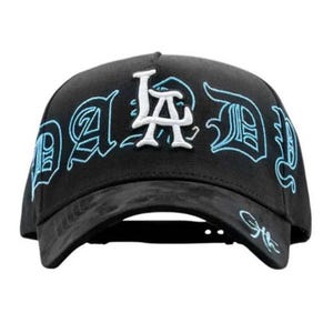 May include: Black baseball cap featuring the word "DAD" in a stylized font, outlined in light blue, and the letters "LA" in white. The cap has a curved peak and a structured crown.