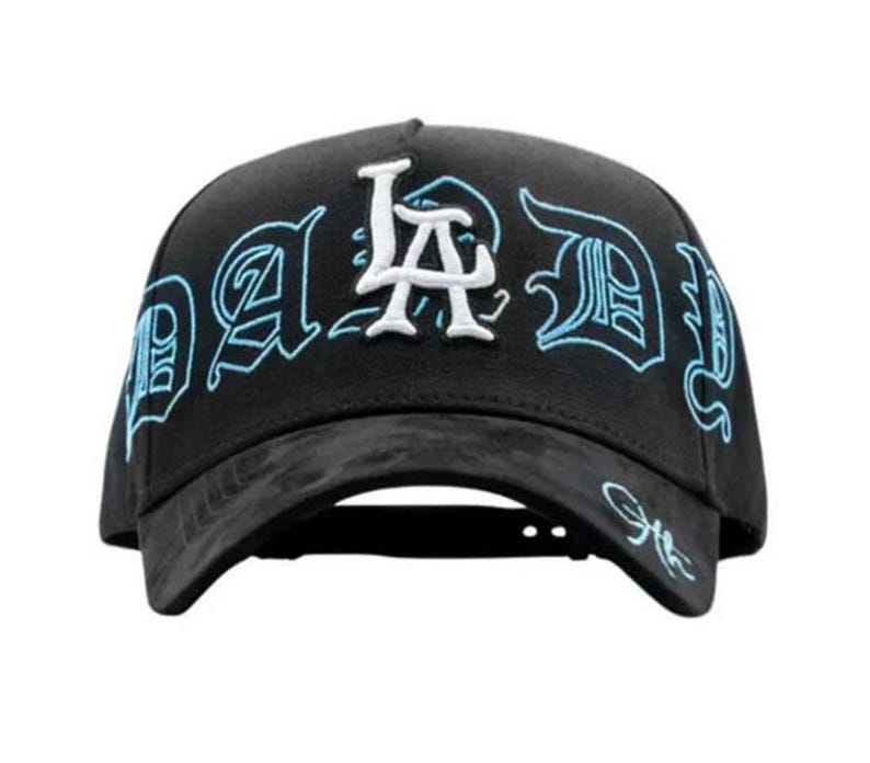 May include: Black baseball cap featuring the word "DAD" in a stylized font, outlined in light blue, and the letters "LA" in white. The cap has a curved peak and a structured crown.