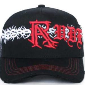 May include: Black baseball cap with red stitching on the brim. The cap features the word "RUDE" in red gothic lettering, with a white thorny design above the word. The cap has a distressed look.
