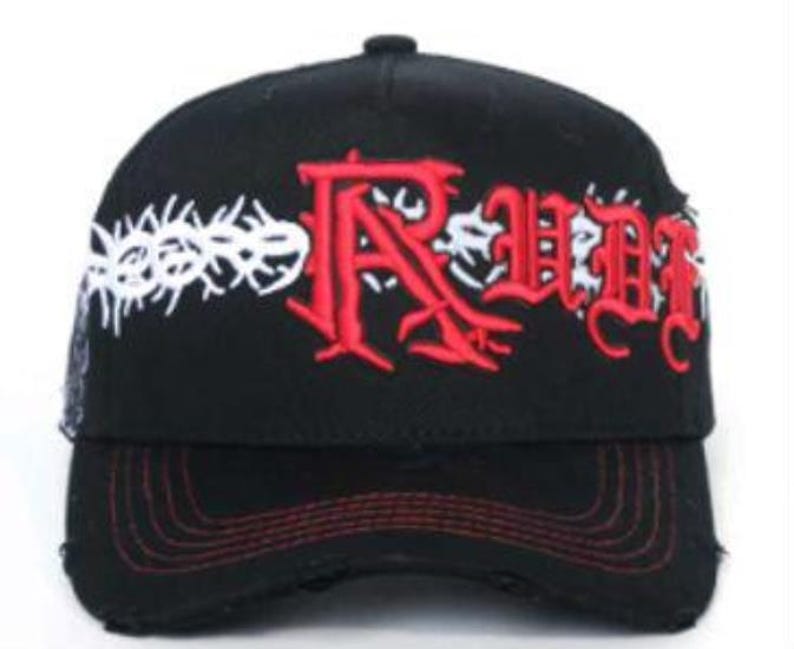 May include: Black baseball cap with red stitching on the brim. The cap features the word "RUDE" in red gothic lettering, with a white thorny design above the word. The cap has a distressed look.