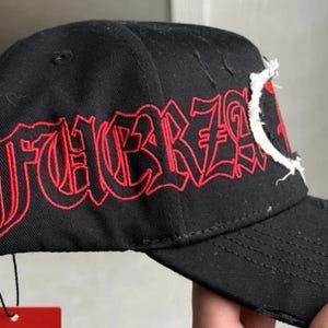May include: A black baseball cap featuring the word FUERZA embroidered in red gothic-style lettering on the side panel. The cap includes distressed fabric details along the brim and a white frayed patch element near the front crown area.