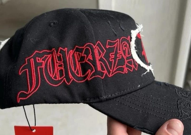 May include: A black baseball cap featuring the word FUERZA embroidered in red gothic-style lettering on the side panel. The cap includes distressed fabric details along the brim and a white frayed patch element near the front crown area.