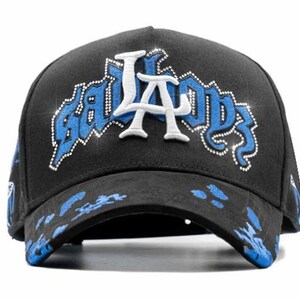 May include: Black baseball cap with the text "BAD BOYS LA" in white and blue, embellished with rhinestones. The brim features blue paint splatters. A stylish accessory for casual wear.