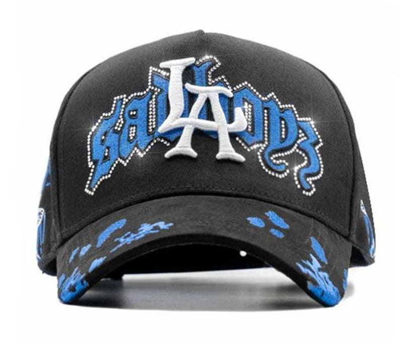 May include: Black baseball cap with the text "BAD BOYS LA" in white and blue, embellished with rhinestones. The brim features blue paint splatters. A stylish accessory for casual wear.