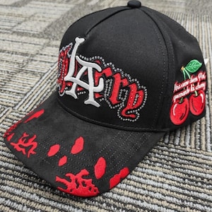 May include: A black baseball cap featuring a white LA logo outlined in silver rhinestones, with red embroidered lettering and a red paint splatter pattern on the brim. The side displays an embroidered cherry graphic with green leaves and text.
