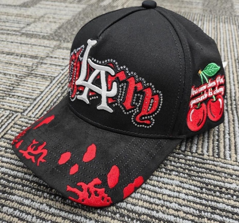 May include: A black baseball cap featuring a white LA logo outlined in silver rhinestones, with red embroidered lettering and a red paint splatter pattern on the brim. The side displays an embroidered cherry graphic with green leaves and text.