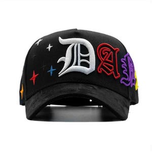 May include: Black baseball cap with embroidered letters and star accents. The cap features a large white "D", a red "A", and a purple letter. Colorful stars and accents are embroidered on the cap.