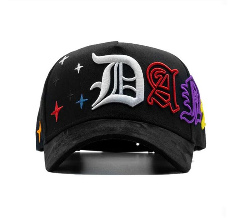 May include: Black baseball cap with embroidered letters and star accents. The cap features a large white "D", a red "A", and a purple letter. Colorful stars and accents are embroidered on the cap.