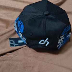 May include: A black baseball cap featuring blue and white embroidered graphic designs on the sides. The back of the cap displays a white embroidered logo with the letters d and h. A small black tag with the text dandy is attached to the side.