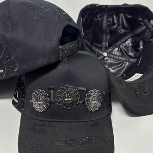 May include: Three black baseball caps featuring a structured crown and flat brim. The front of the center cap displays three circular flower-shaped metal emblems with black stones. The interior lining is black satin with repeating text.