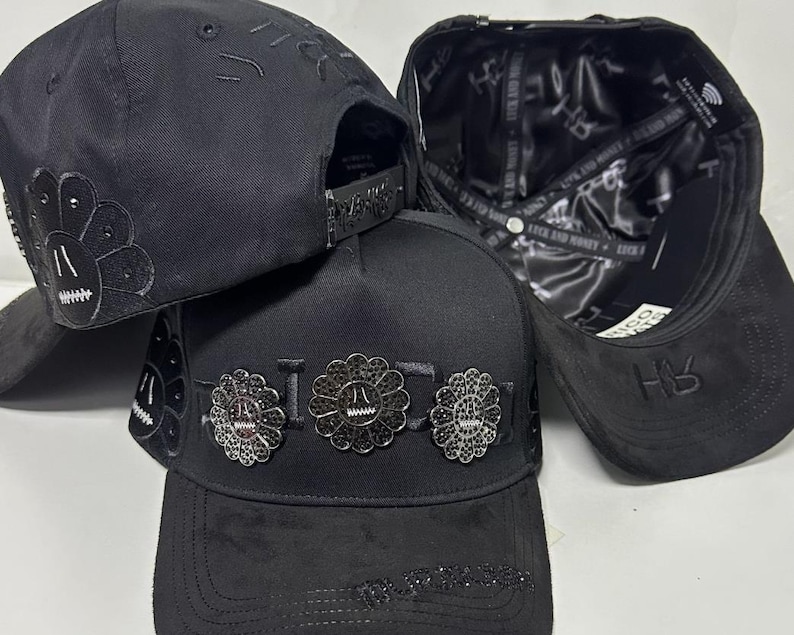 May include: Three black baseball caps featuring a structured crown and flat brim. The front of the center cap displays three circular flower-shaped metal emblems with black stones. The interior lining is black satin with repeating text.
